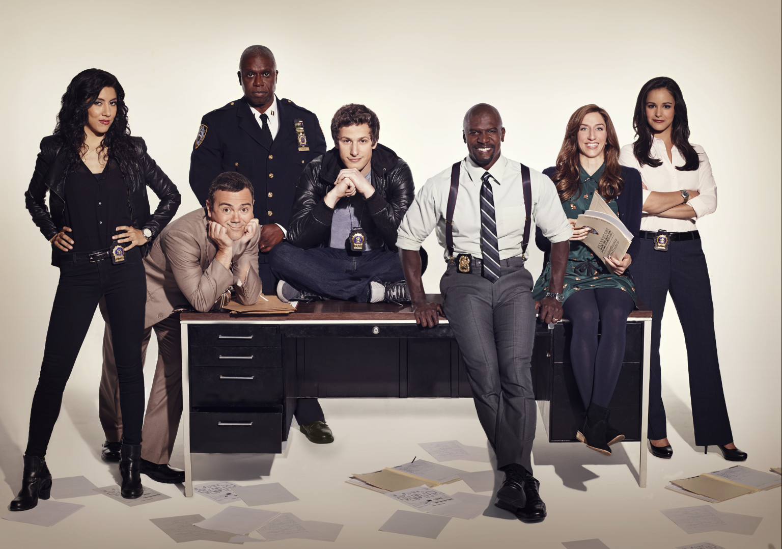 The original Brooklyn Nine-Nine cast around a precinct desk in 2013 publicity photography: Rosa, Holt, Boyle, Jake, Terry, Gina, and Amy.